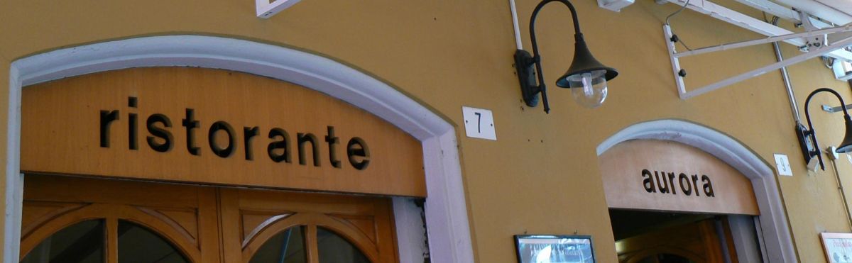 Le insegne del Ristorante Aurora: le parole ristorante e aurora in lettere nere minuscole sugli architravi dei portoni, muro color ocra
